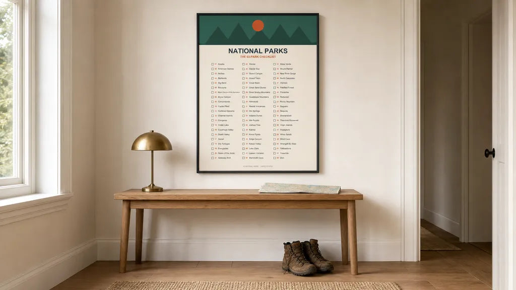 The RaZits National Parks 63-Park Checklist poster in a thin black frame on a hallway wall above an oak bench with a brass lamp, a folded map and worn hiking boots beneath