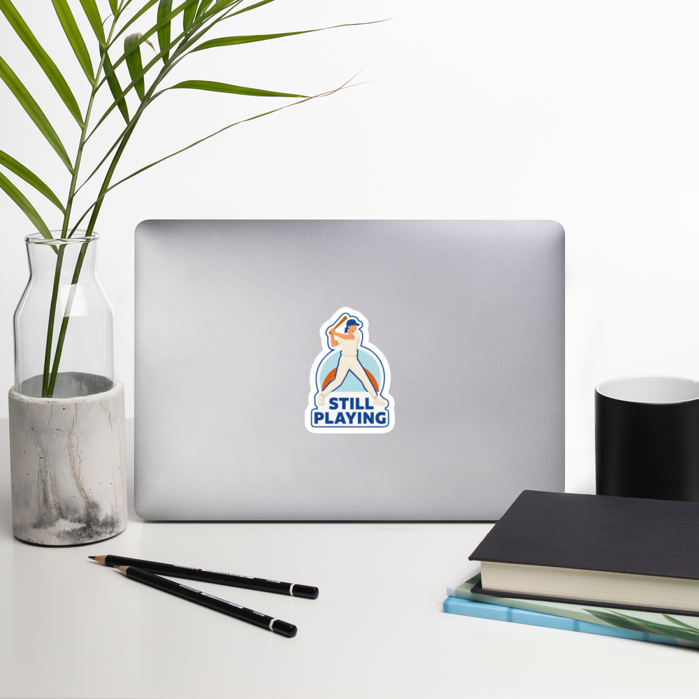STILL PLAYING 4 inch baseball sticker on a laptop in a front-facing indoor desk scene with books, a mug and a plant. Styling props are not included.
