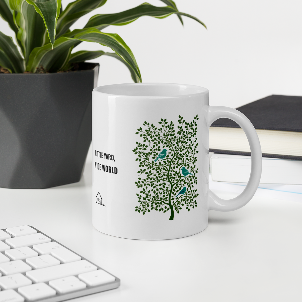 11 oz backyard bird mug on a desk beside a plant and keyboard, showing LITTLE YARD, WIDE WORLD beside a leafy tree with three teal birds