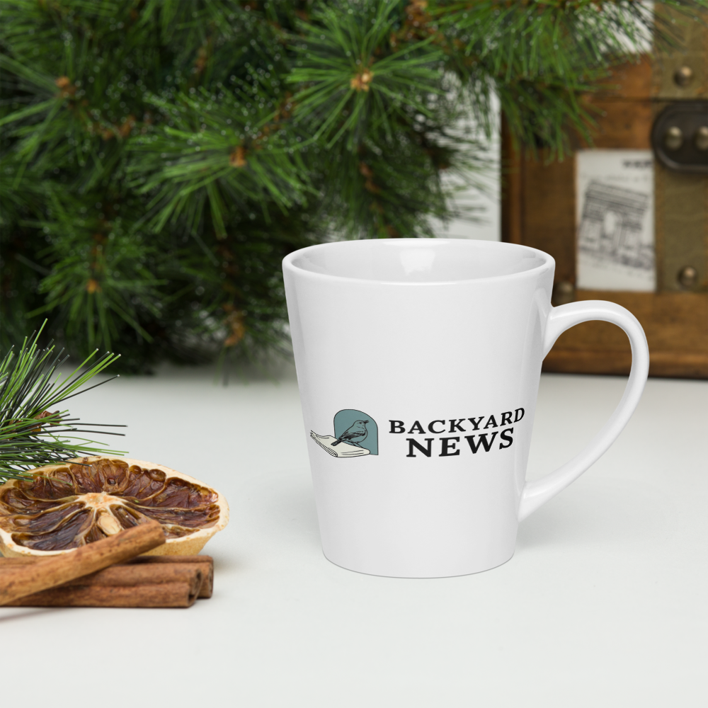 Bird coffee mug on a table with pine branches, dried orange and cinnamon, showing a songbird on a folded newspaper beside BACKYARD NEWS