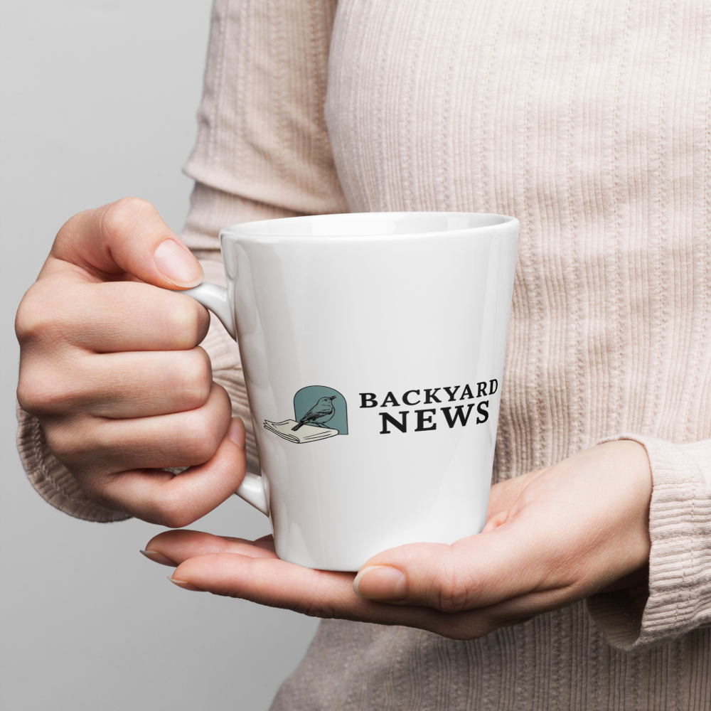 Hands holding the 12 oz bird coffee mug, showing a songbird on a folded newspaper beside BACKYARD NEWS