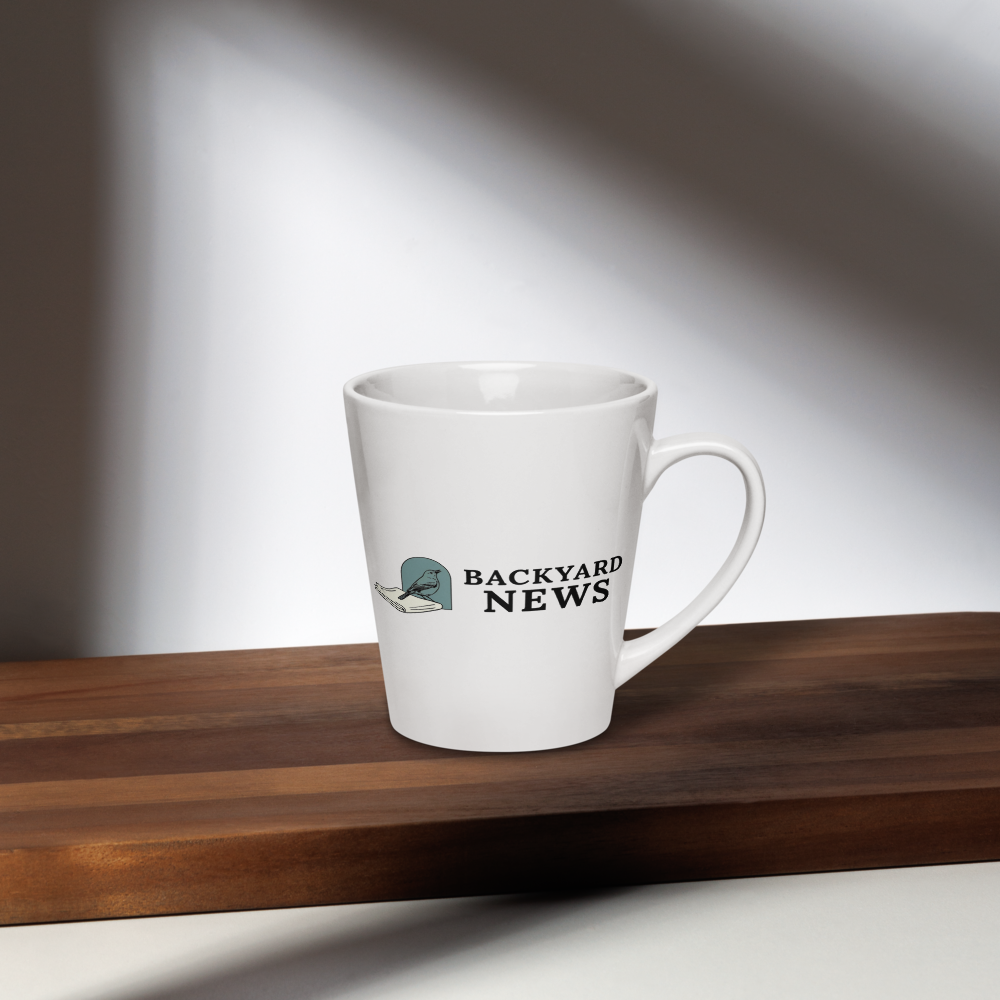 Bird coffee mug on a dark wood shelf in window light, showing a songbird on a folded newspaper beside BACKYARD NEWS