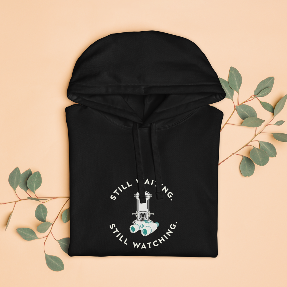 The black bird hoodie folded with eucalyptus leaves, showing STILL WAITING. STILL WATCHING. around a feeder and binoculars