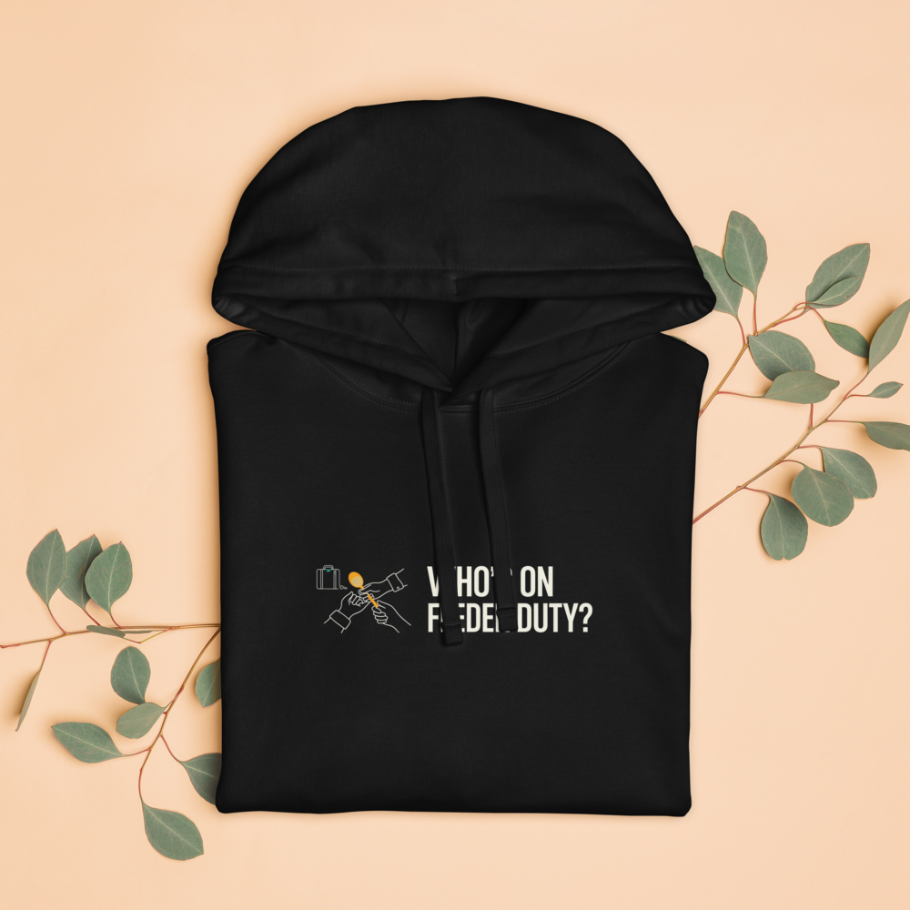 The black bird lover hoodie folded with eucalyptus leaves, showing two hands passing a seed scoop beside WHO'S ON FEEDER DUTY?