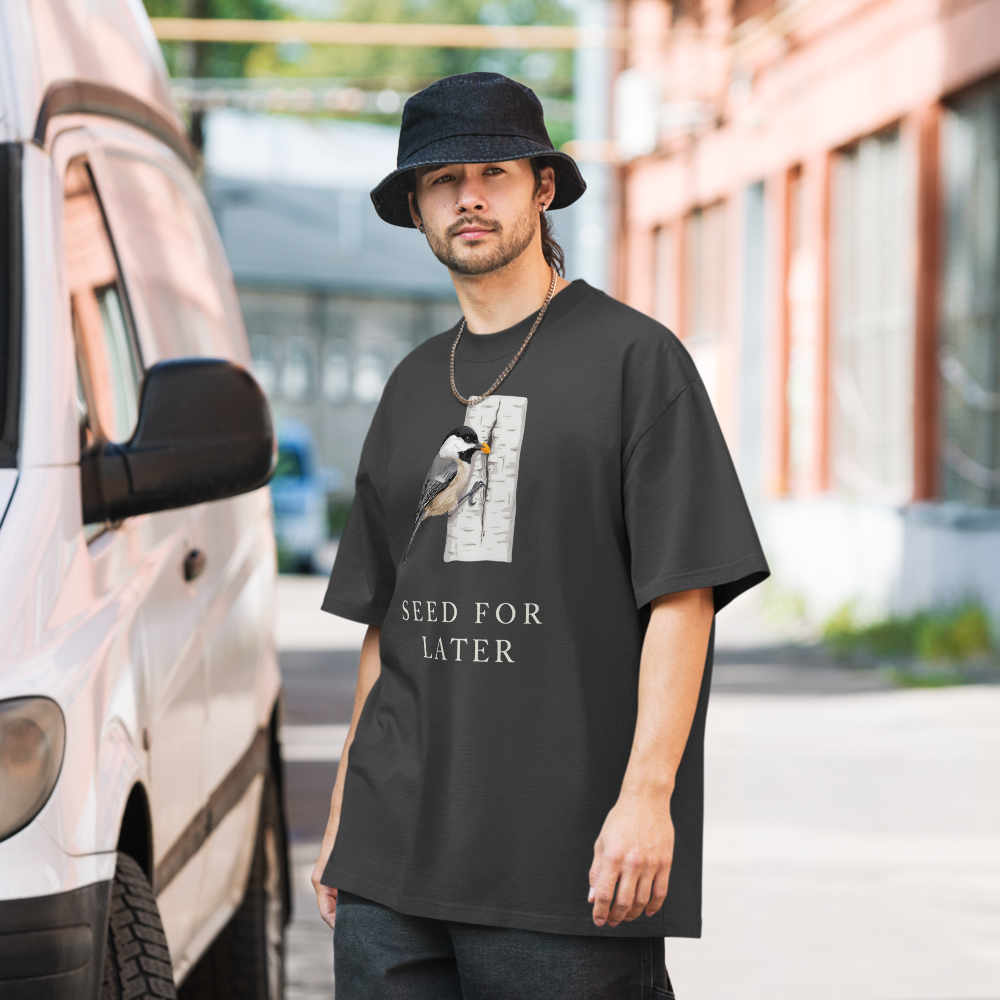 Man in the faded black bird t shirt on a city street, showing a chickadee tucking a seed into birch bark above SEED FOR LATER