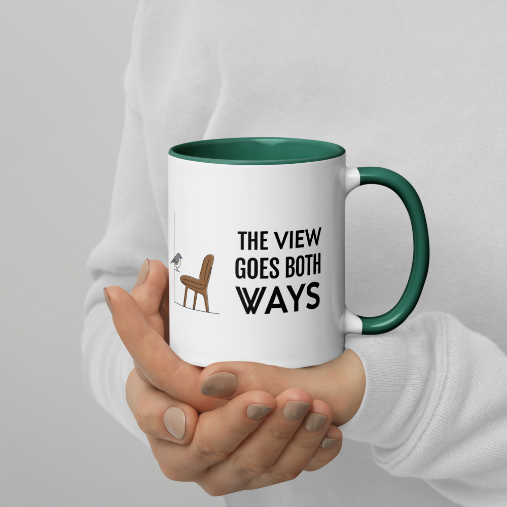 Hands holding the bird watching mug with dark green inside, showing THE VIEW GOES BOTH WAYS beside a bird on the sill and an empty chair