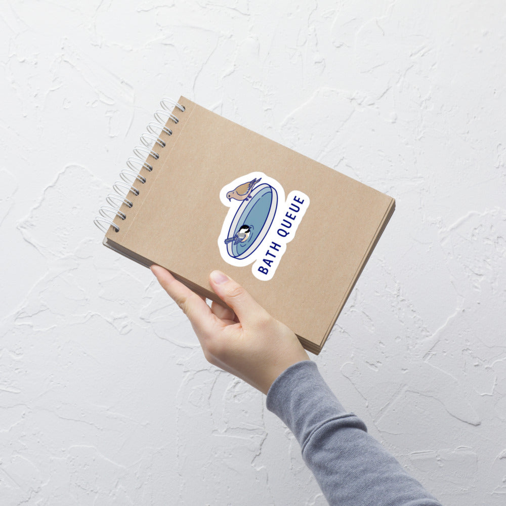 Hand holding a kraft notebook with the Bath Queue bird sticker on the cover