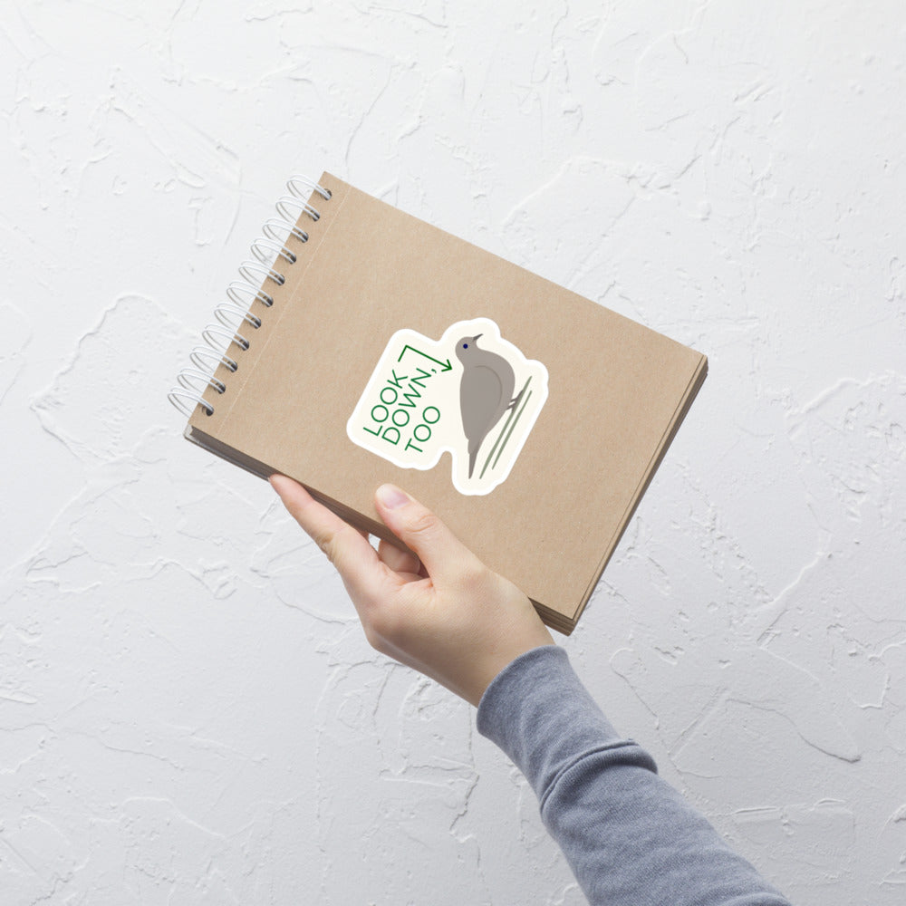 Hand holding a kraft notebook with the Look Down, Too dove sticker on the cover