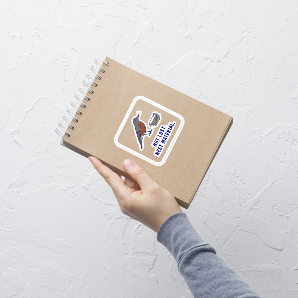 Hand holding a kraft notebook with the Not Lost. Nest Material. robin sticker on the cover