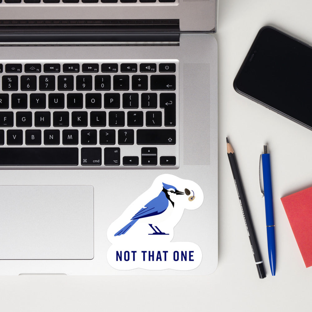 Not That One blue jay sticker on a laptop next to pens and a phone