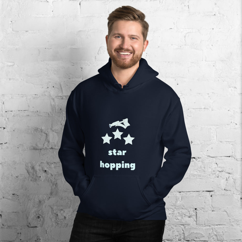 Smiling man wearing the navy Star Hopping hoodie with a telescope, three stars and star hopping lettering, front view by a white brick wall