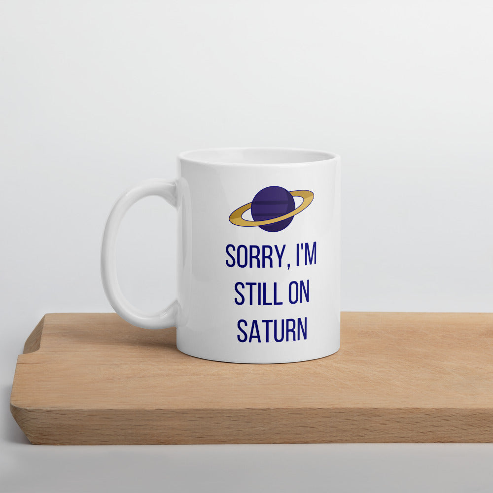 Sorry, I'm Still on Saturn mug standing on a wooden board, purple planet with a gold ring above the navy lettering
