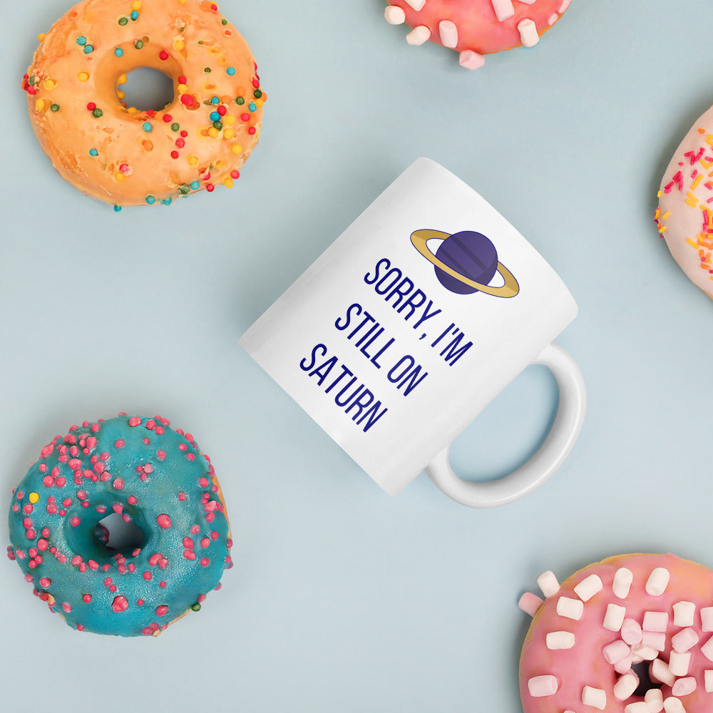 Sorry, I'm Still on Saturn astronomy mug lying among sprinkled donuts on a pale blue table