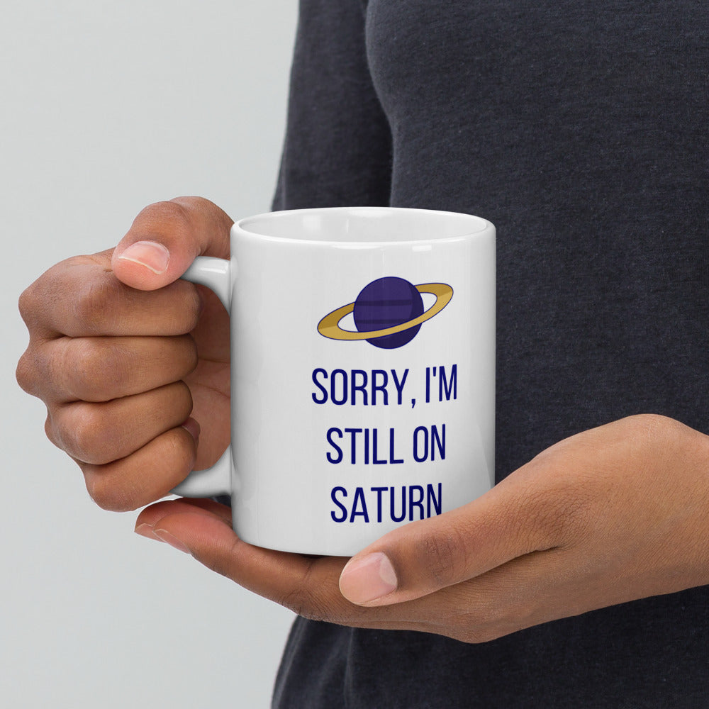 Hand holding the Saturn mug by its handle, SORRY, I'M STILL ON SATURN and the gold-ringed planet facing out