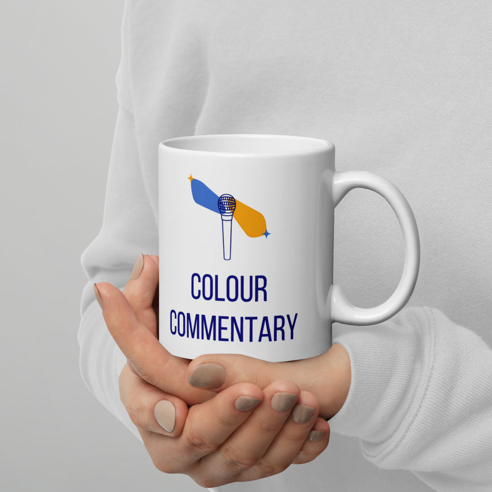 Hands in a white sweater cradling the 11 oz double star mug, Colour Commentary lettering and blue and gold streaks facing out