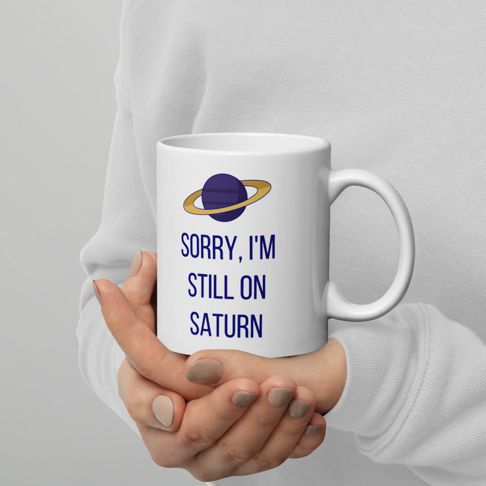 Hands in a white sweater cradling the 11 oz Saturn mug, the purple planet with its gold ring and the navy lettering facing out
