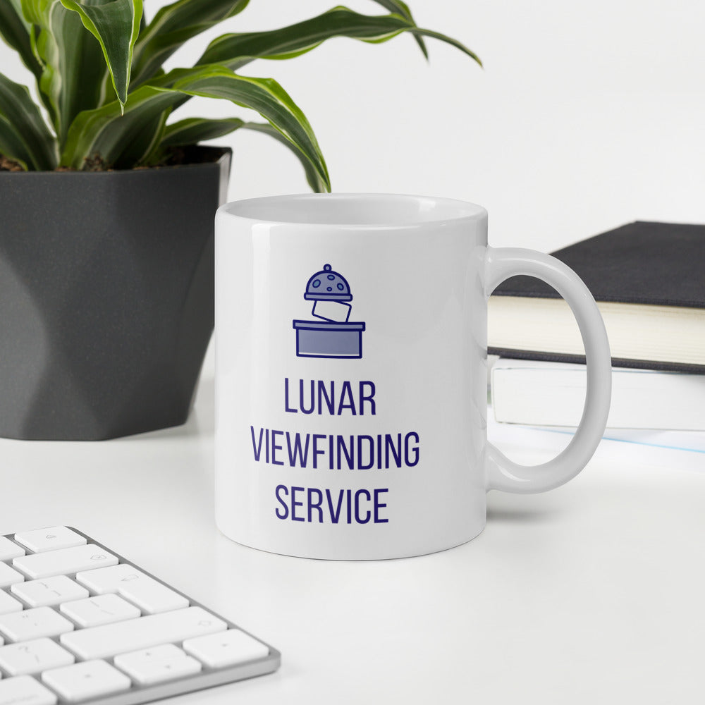 Lunar Viewfinding Service mug on a white desk beside a keyboard, notebook and potted plant