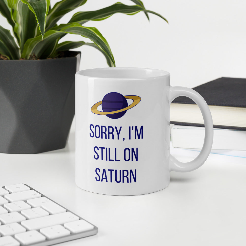 Sorry, I'm Still on Saturn astronomy mug on a white desk beside a keyboard, a plant and a stack of books