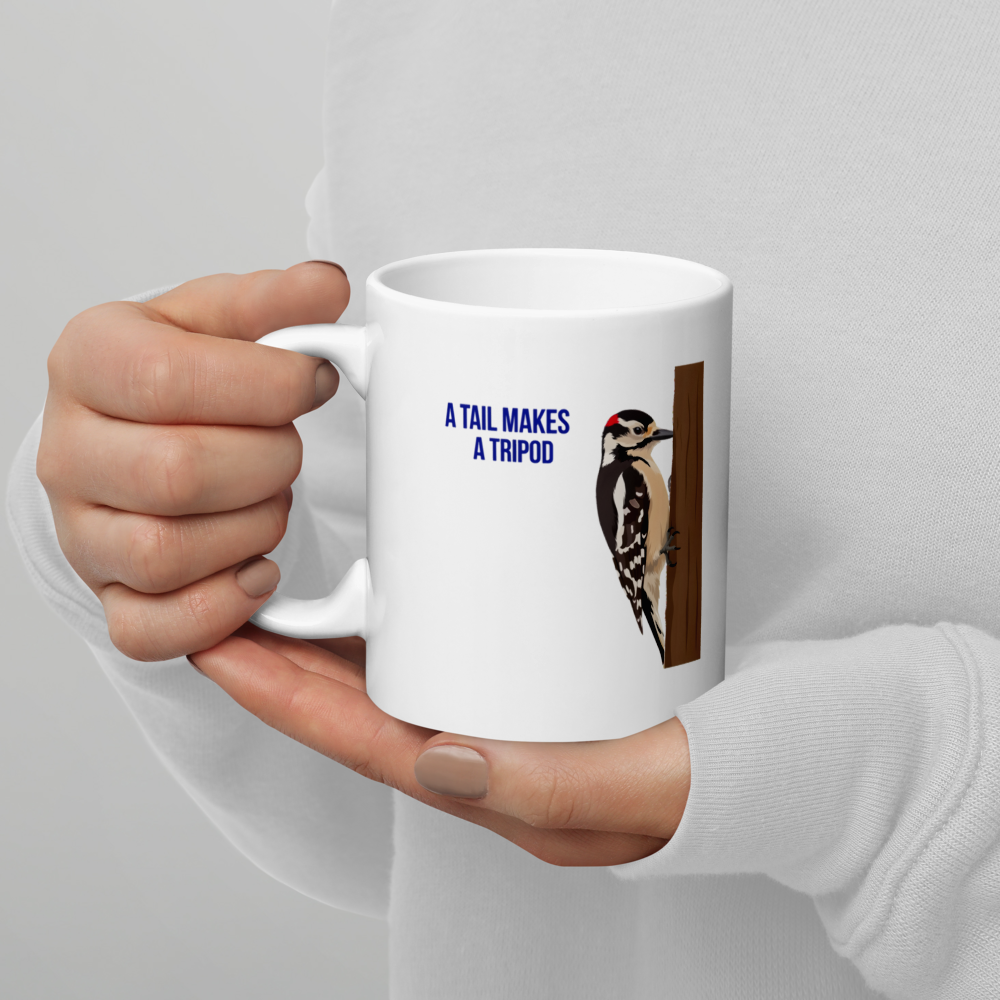 Hands holding the 11 oz woodpecker mug, A Tail Makes a Tripod lettering and Downy Woodpecker on the trunk facing out