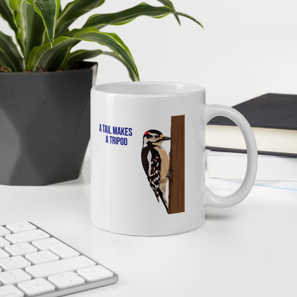 Woodpecker mug on an office desk beside a plant and keyboard, showing the A Tail Makes a Tripod Downy Woodpecker design